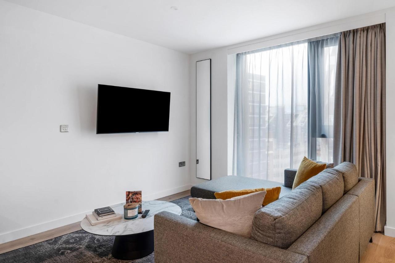 Citysuites 2 Aparthotel マンチェスター エクステリア 写真 The image shows a modern living room with a minimalist design. There is a large flat-screen TV mounted on the white wall. A comfortable gray sofa with textured cushions occupies the center of the room, complemented by a round coffee table in front of