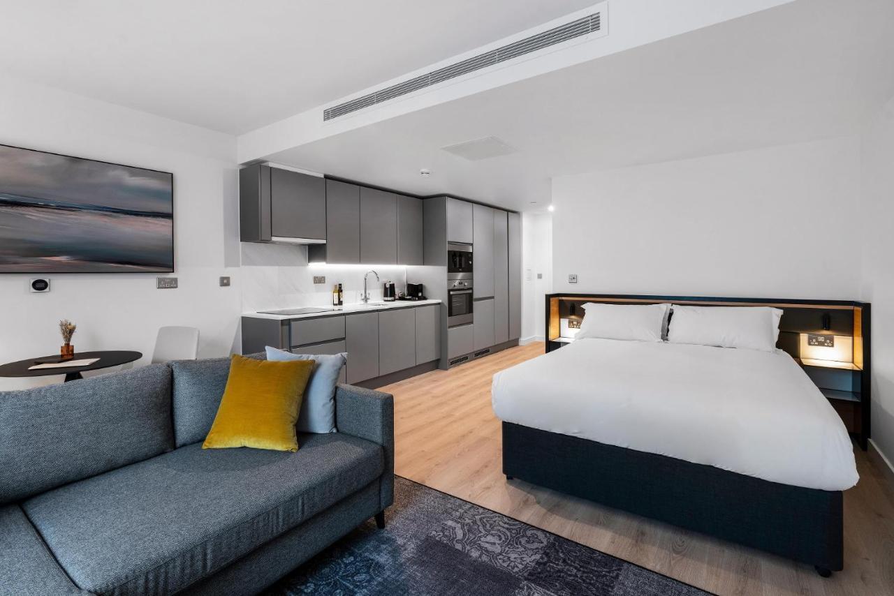Citysuites 2 Aparthotel マンチェスター エクステリア 写真 The photo shows a modern studio apartment. On one side, there is a comfortable gray sofa adorned with a yellow cushion. Nearby, a round table is placed, likely intended for dining or casual sitting. 

The kitchen area features sleek gray cabinetry wi