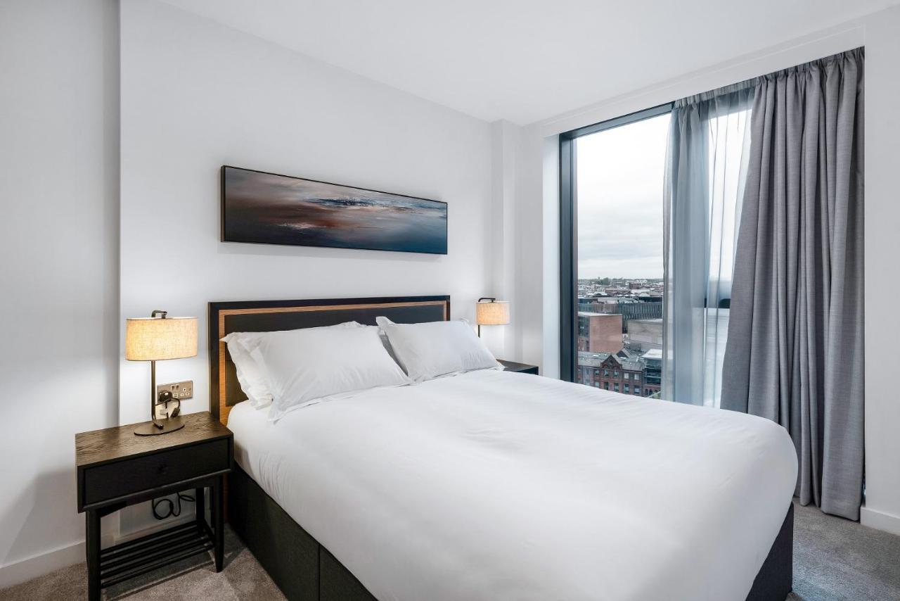 Citysuites 2 Aparthotel マンチェスター エクステリア 写真 The image shows a modern bedroom featuring a large bed with a white duvet and two pillows. On either side of the bed are small nightstands with lamps. A large window occupies one wall, draped with light gray curtains, allowing natural light to enter 