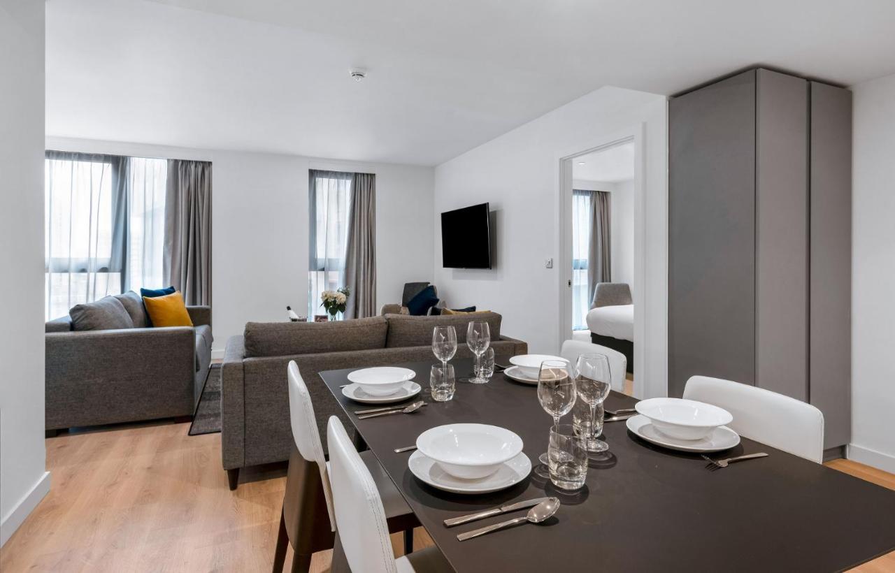 Citysuites 2 Aparthotel マンチェスター エクステリア 写真 The photo shows a modern living space with an open layout. In the foreground, there is a dining table set for a meal, featuring multiple plates, glasses, and cutlery arranged neatly. In the background, there is a comfortable-looking gray sofa with a 