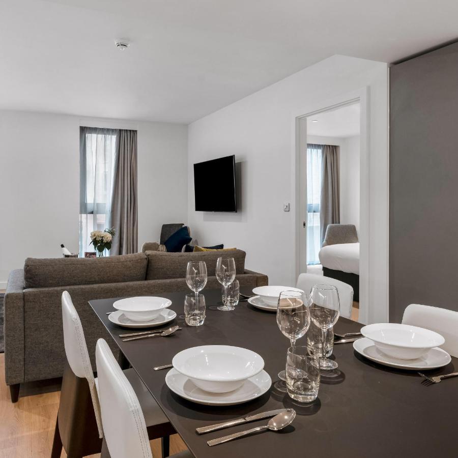 Citysuites 2 Aparthotel マンチェスター エクステリア 写真 The photo shows a modern, well-lit living space that features a dining area and a sitting area. In the foreground, there is a dining table set for six, with white plates and crystal glasses arranged neatly. The table is surrounded by white chairs. 

