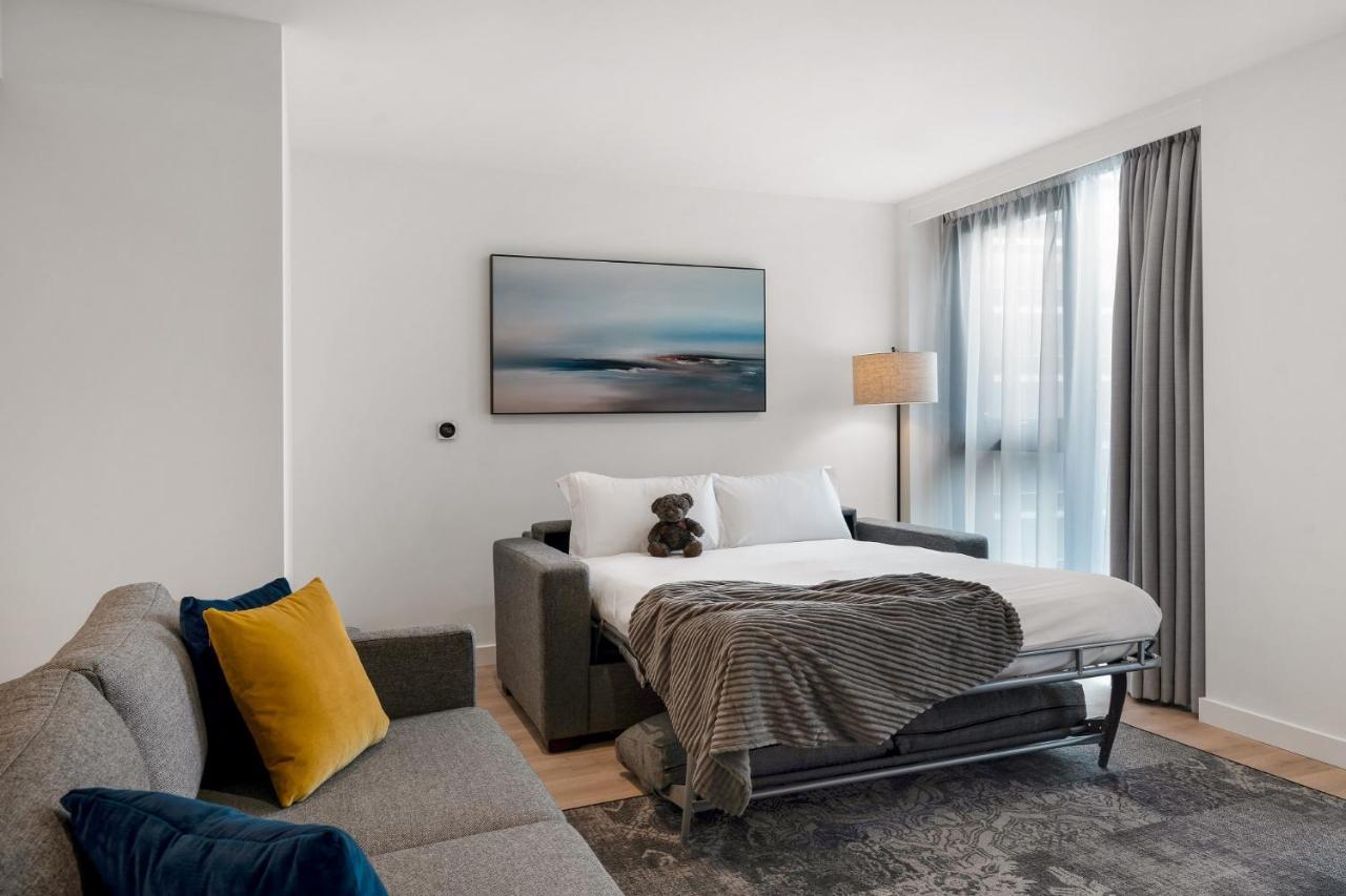 Citysuites 2 Aparthotel マンチェスター エクステリア 写真 The photo shows a modern and minimalistic guest room. There is a sofa on the left side, adorned with a colorful cushion. In the middle is a bed that appears to be a pull-out style, featuring white bedding and a gray throw blanket. A stuffed toy is pl