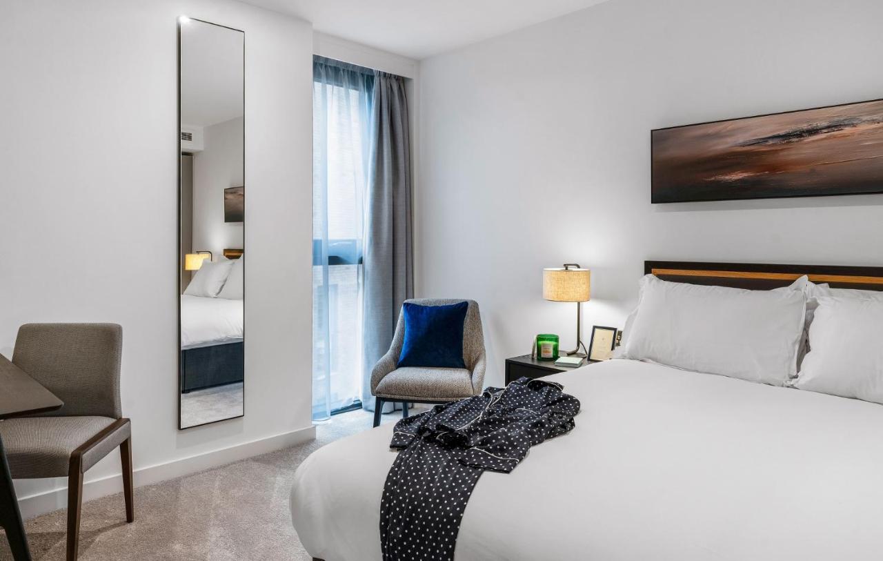 Citysuites 2 Aparthotel マンチェスター エクステリア 写真 The photo shows a modern hotel room. It features a neatly made bed with white linens and a dark-colored throw draped over it. There is a small bedside table with a lamp next to the bed. A comfortable chair in a light color is positioned near the wind