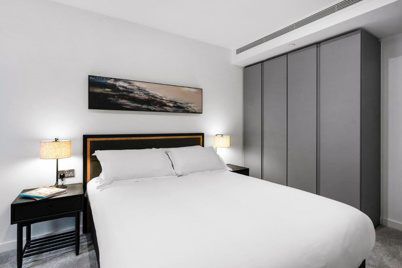 Citysuites 2 Aparthotel マンチェスター エクステリア 写真 The photo shows a modern bedroom designed with a minimalist aesthetic. The focal point is a large bed with white bedding, positioned against a wall. On either side of the bed, there are black nightstands topped with lamps, providing a warm glow. Abov