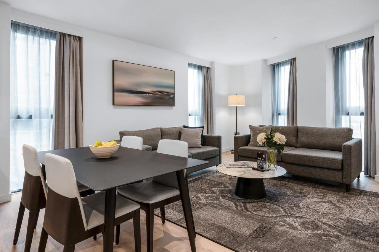 Citysuites 2 Aparthotel マンチェスター エクステリア 写真 The photo depicts a modern and stylish interior of a living space. On the left, there is a dining table with four chairs, featuring a bowl of fruits on the table. In the background, there's a cozy seating area with a gray sofa and a smaller round cof