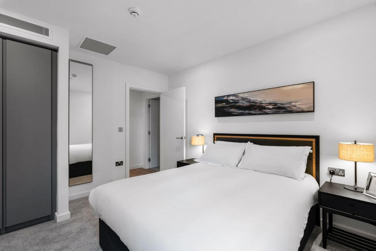 Citysuites 2 Aparthotel マンチェスター エクステリア 写真 The photo shows a modern bedroom featuring a large double bed with white bedding. There are two bedside tables with lamps on either side of the bed. The walls are painted white, and there's a piece of artwork above the bed that depicts a landscape. A