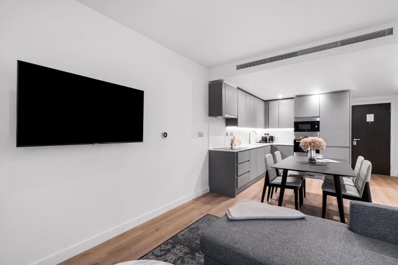 Citysuites 2 Aparthotel マンチェスター エクステリア 写真 The photo shows a modern, minimalist living space. On the left, there is a large flat-screen TV mounted on the wall. To the right, a kitchen area features sleek gray cabinets and appliances, including a built-in microwave. In front of the kitchen, th