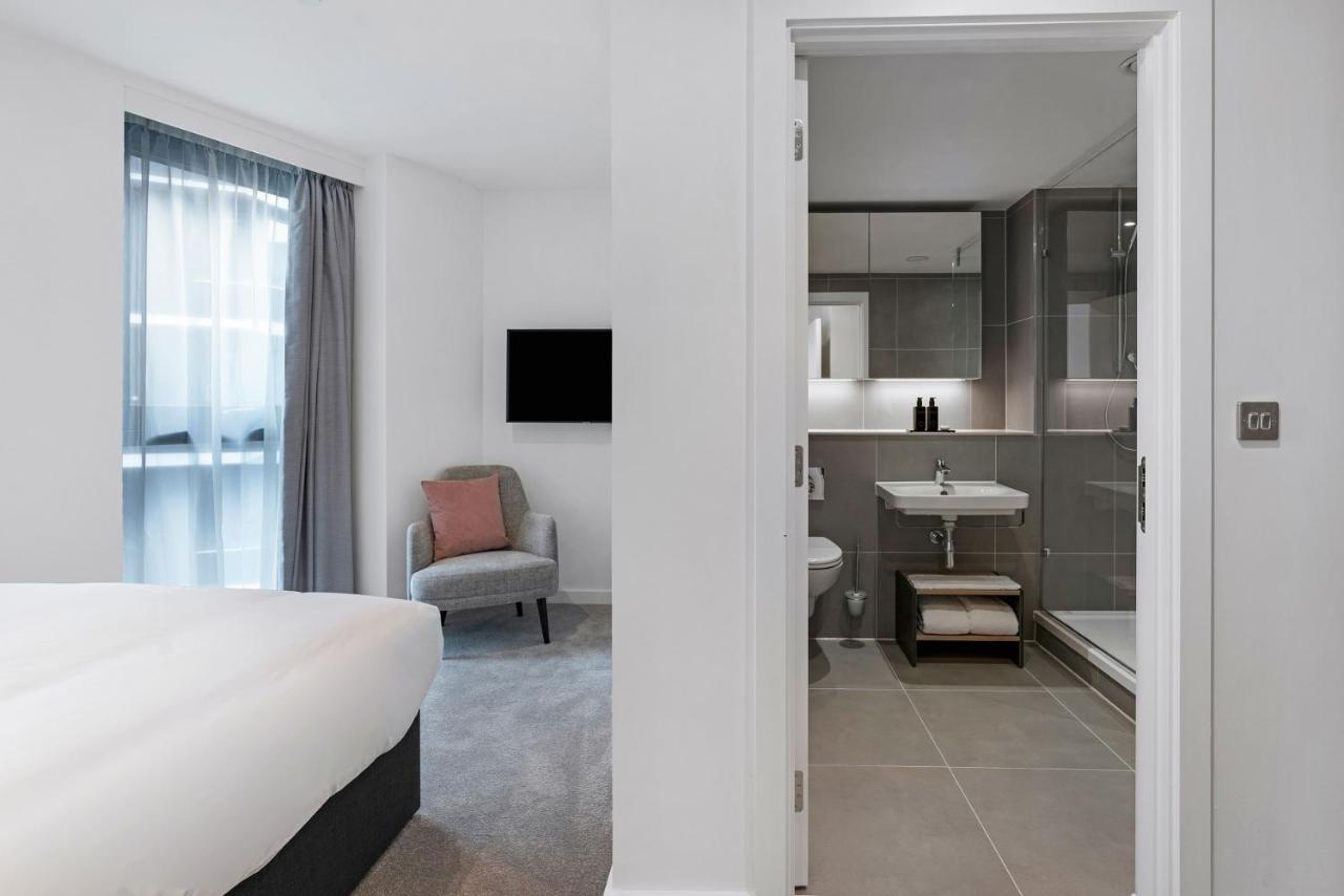 Citysuites 2 Aparthotel マンチェスター エクステリア 写真 The photo shows a modern hotel room and bathroom setup. On the left, there is a bed with white bedding and a small sitting area that includes a gray chair and a pillow in a subtle pink shade. A flat-screen TV is mounted on the wall, providing enterta