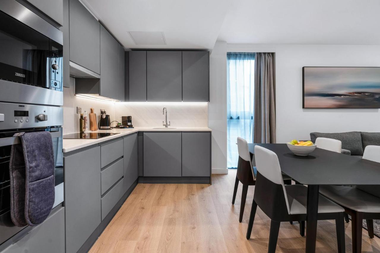 Citysuites 2 Aparthotel マンチェスター エクステリア 写真 The photo shows a modern kitchen and dining area. The kitchen features sleek gray cabinetry, a white countertop, and built-in appliances such as an oven. There is a sink and some kitchen utensils displayed. The adjacent dining area contains a black t