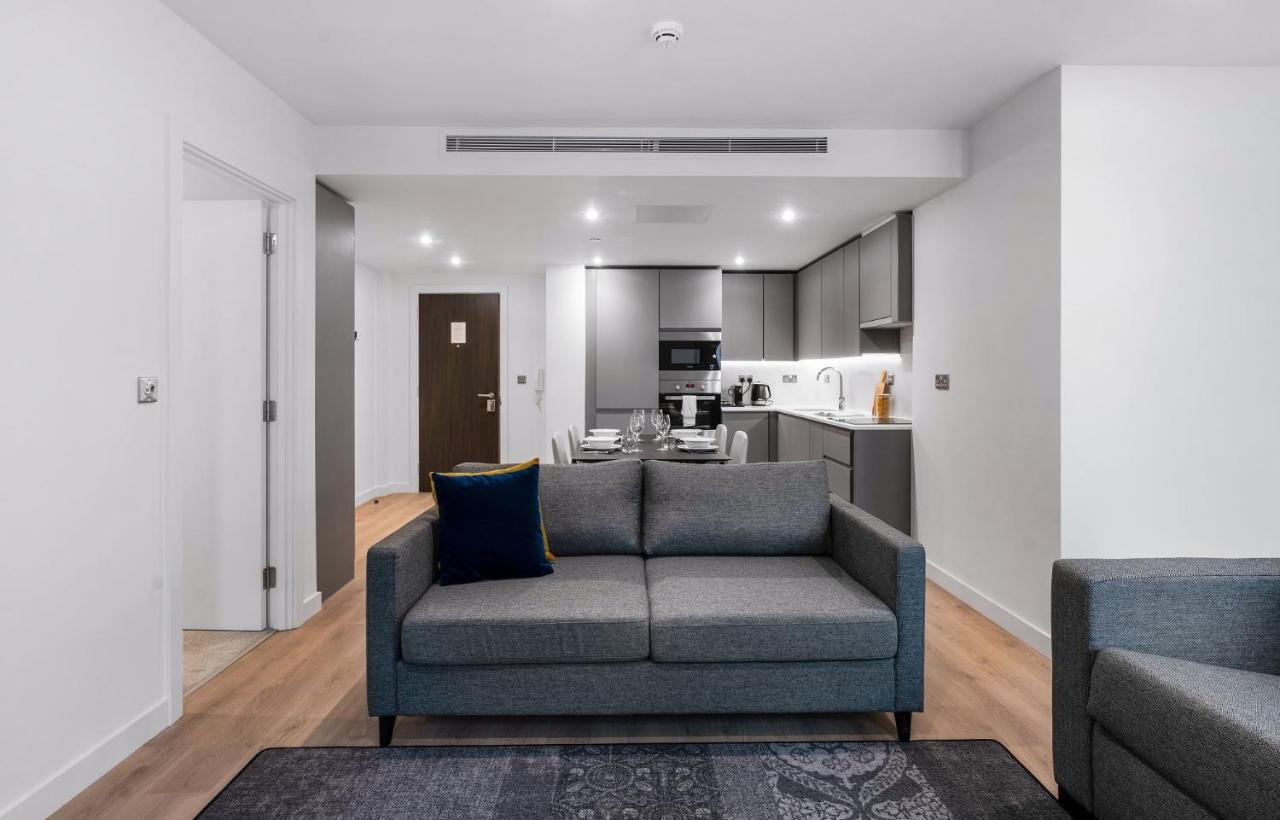 Citysuites 2 Aparthotel マンチェスター エクステリア 写真 The photo shows a modern interior space, likely a living area or apartment. In the foreground, there is a gray sofa with a blue cushion positioned on a textured rug. To the back, there is an open kitchen with sleek cabinetry and appliances, visible a