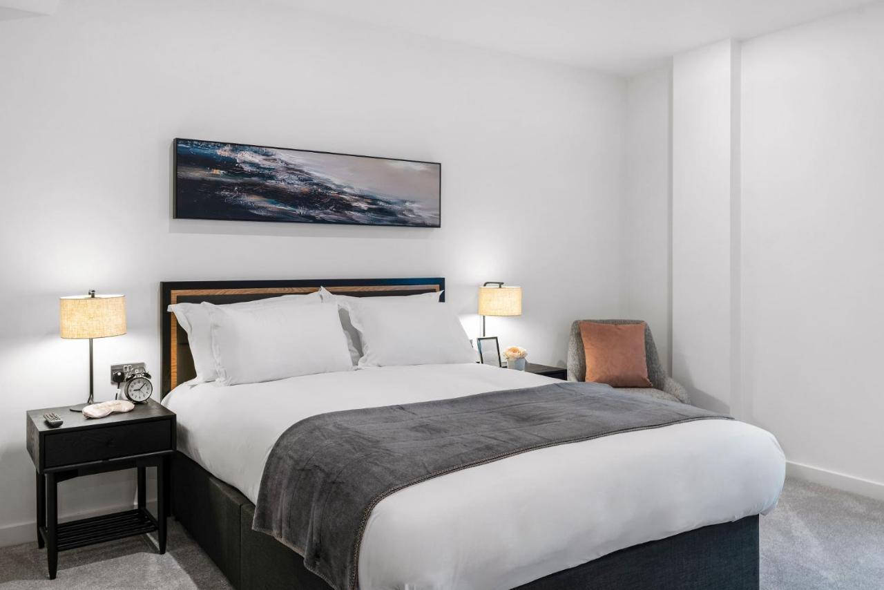 Citysuites 2 Aparthotel マンチェスター エクステリア 写真 The photo shows a modern bedroom featuring a large bed with crisp white linens and a grey throw blanket. On either side of the bed, there are bedside tables with lamps. A piece of abstract art hangs above the headboard, and there is a chair in a corn