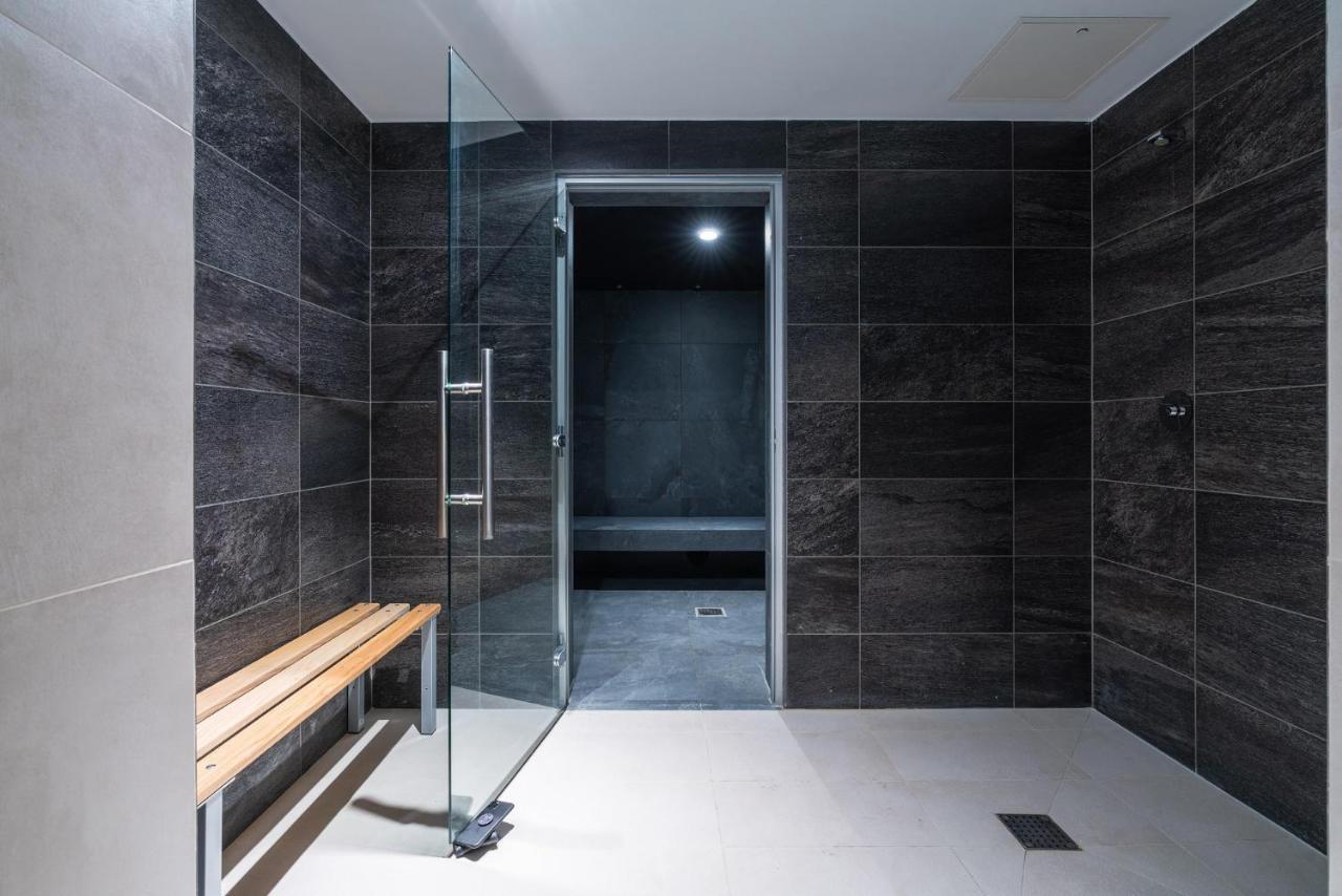 Citysuites 2 Aparthotel マンチェスター エクステリア 写真 The photo shows a modern, minimalist shower area. The space is predominantly tiled with dark gray tiles on the walls, giving it a sleek and sophisticated look. There is a glass door that leads to another room, and a wooden bench is positioned against