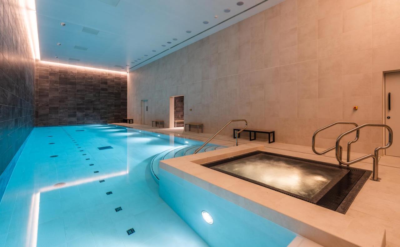Citysuites 2 Aparthotel マンチェスター エクステリア 写真 The photo shows an indoor swimming pool area. The pool is a bright blue color and features smooth, tiled surfaces. On one side, there is a hot tub area with steps leading into it. The walls are made of light-colored stone, and there is ambient lighti