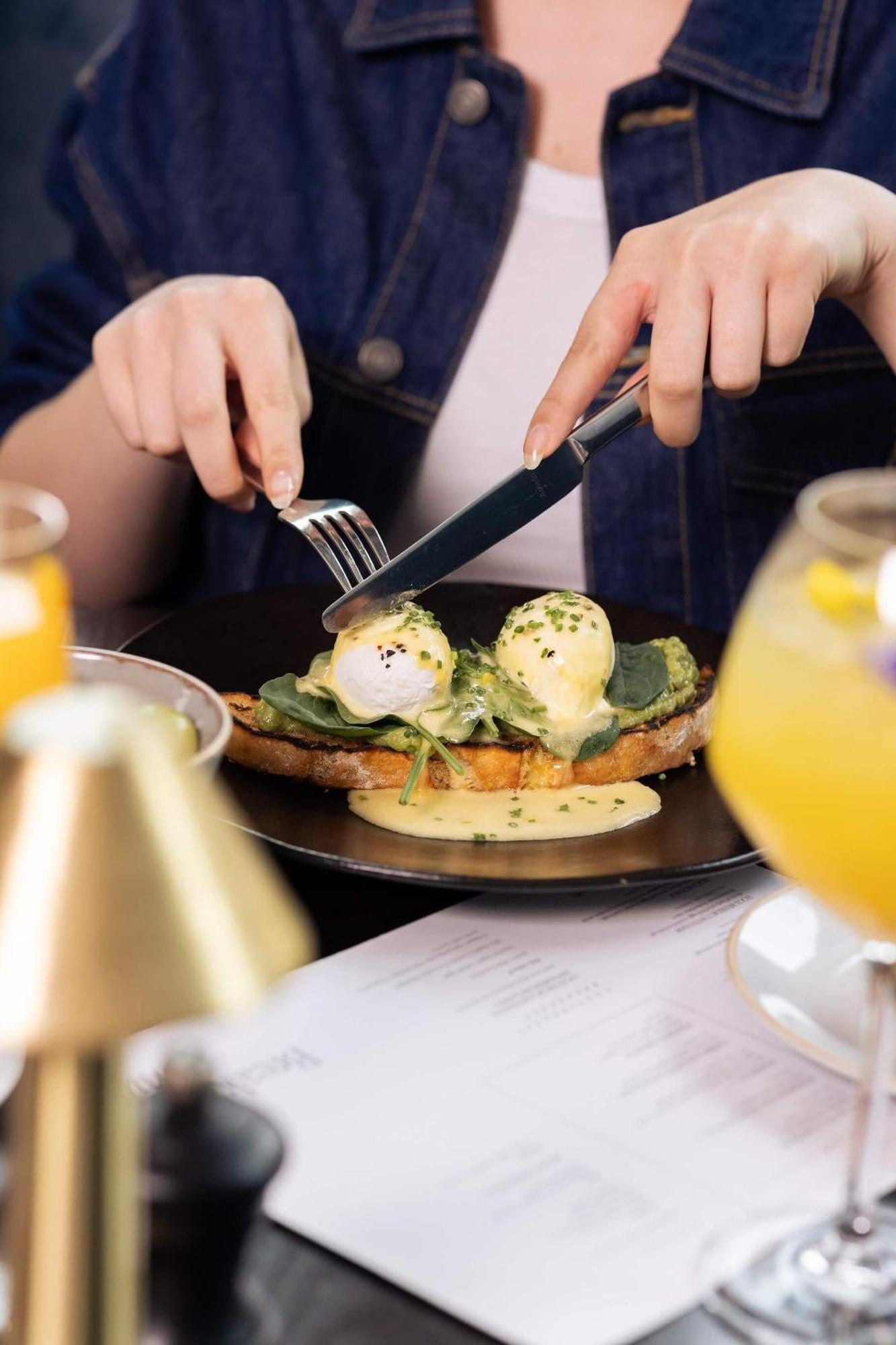 Citysuites 2 Aparthotel マンチェスター エクステリア 写真 The photo shows a person preparing to eat a dish of eggs Benedict. They are holding a knife and fork, ready to cut into the meal, which features poached eggs on top of a slice of toast, accompanied by spinach and a creamy hollandaise sauce drizzled o