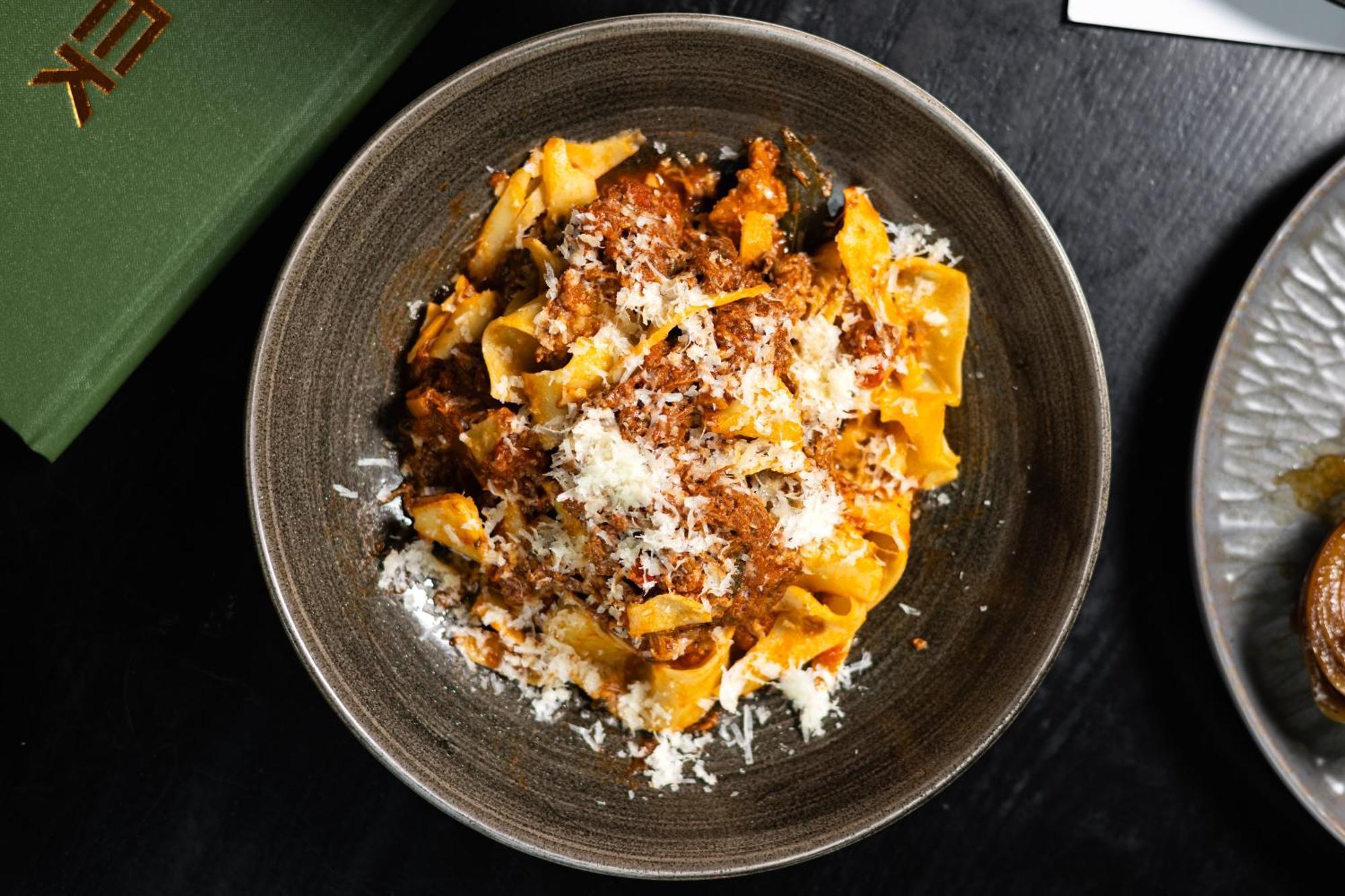 Citysuites 2 Aparthotel マンチェスター エクステリア 写真 The photo shows a bowl of pasta featuring wide, ribbon-like noodles, likely pappardelle, fully coated in a rich meat sauce. The dish is garnished with a generous sprinkling of grated cheese, adding a creamy texture to the presentation. The dark bowl 