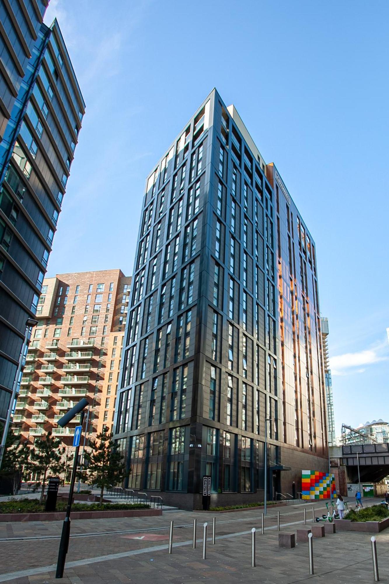 Citysuites 2 Aparthotel マンチェスター エクステリア 写真 The photo shows a modern high-rise building with a sleek, glass façade. It is set against a blue sky, indicating a clear day. The building appears to have multiple floors with large windows that reflect light. Nearby, there are additional buildings, 